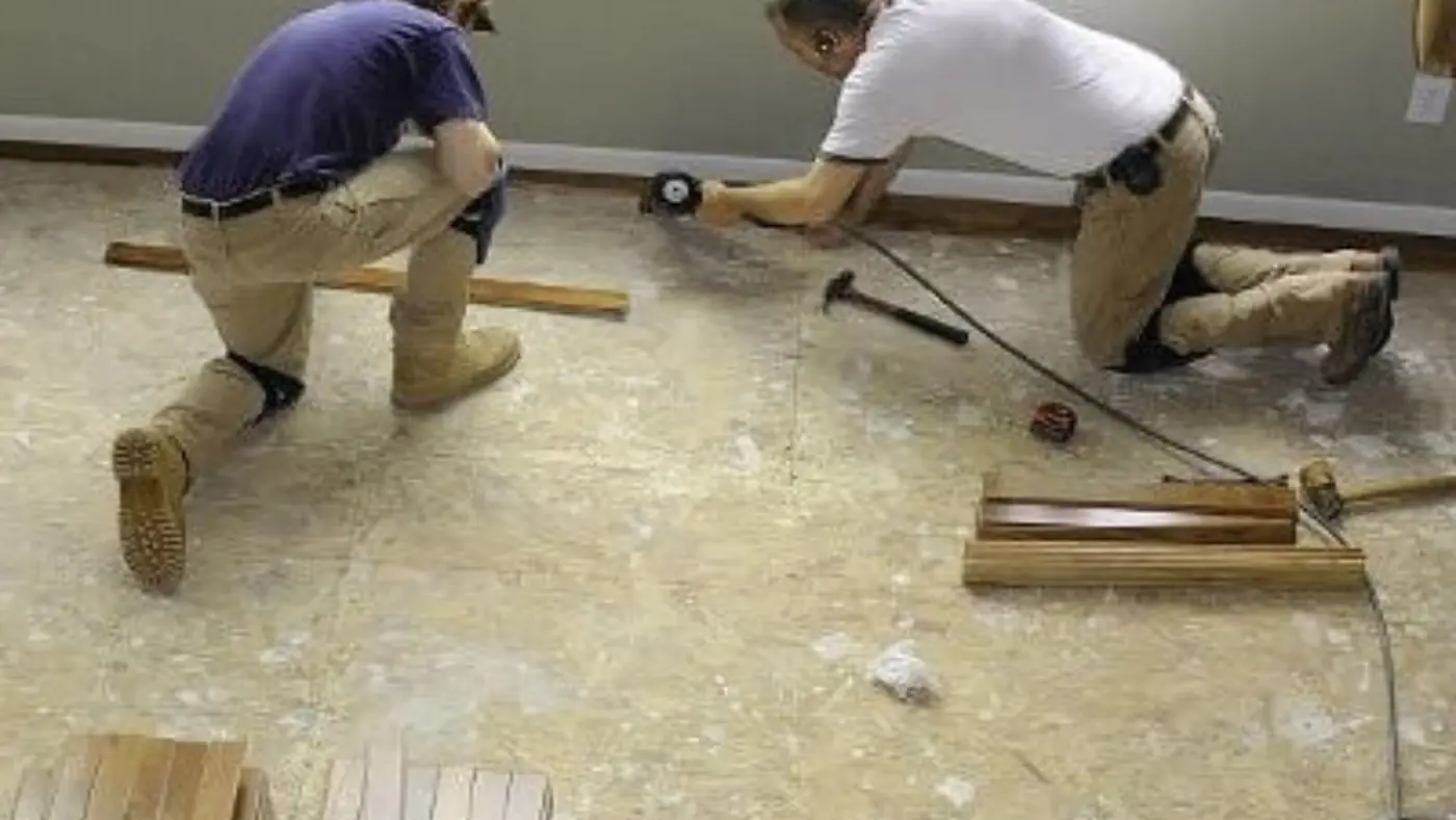 Two workers kneeling on a subfloor measuring and fitting new flooring boards